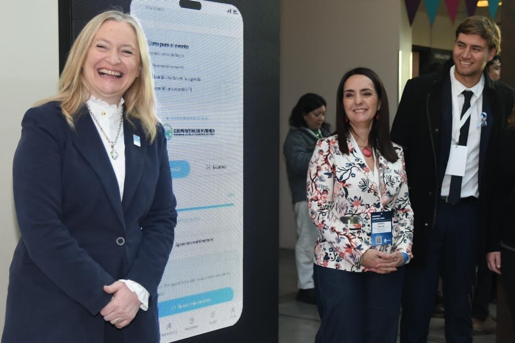 Dos organizadoras junto a una pantalla que muestra el portal del evento en un teléfono; una lleva la credencial con el código QR